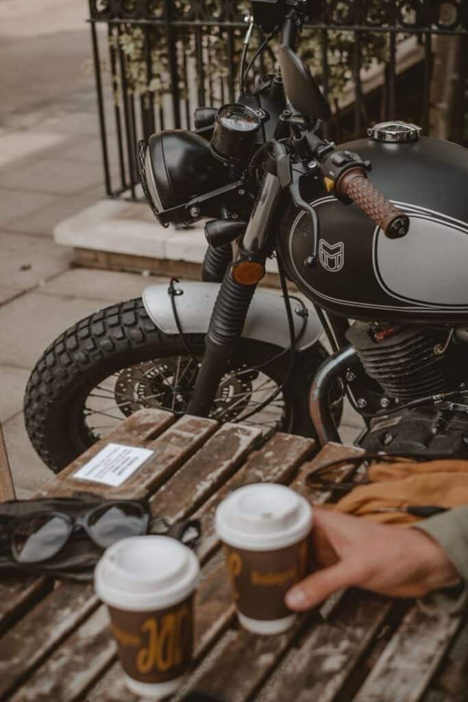 Motorcycle parked by outdoor cafe table with coffee cups and sunglasses on wooden surface.