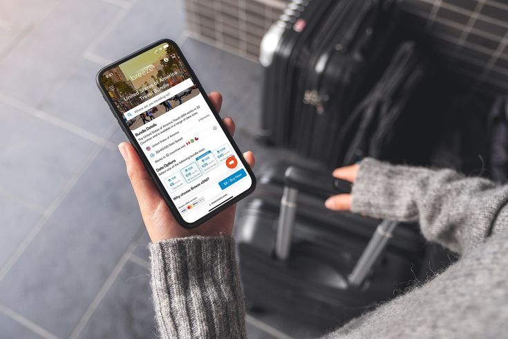 Person using smartphone travel app while standing near suitcase, planning trip.