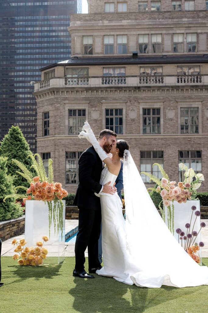 Bride and groom share a kiss during outdoor wedding ceremony with elegant floral decorations and city backdrop.