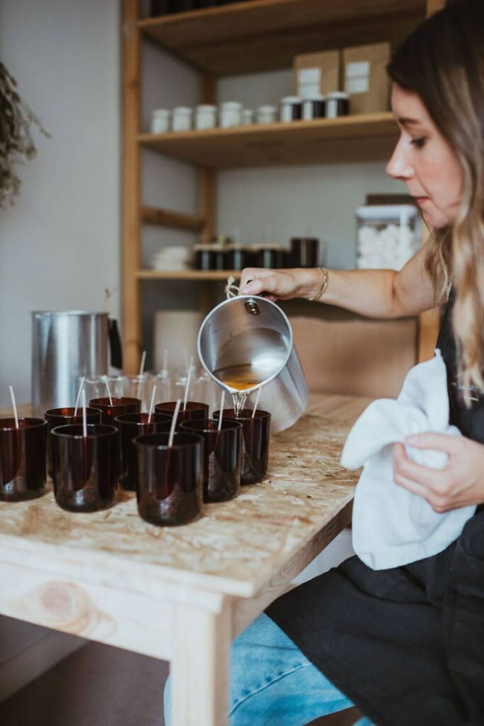 Person pouring wax into candle molds in a craft workshop, surrounded by materials on a wooden table.