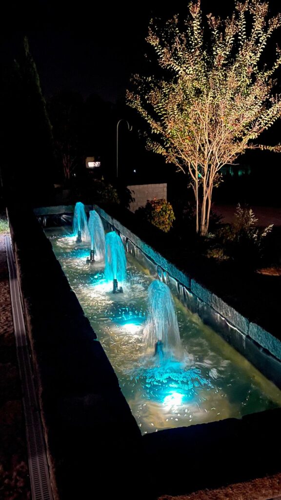 Illuminated night fountain with cascading blue-lit water and tree in the background.