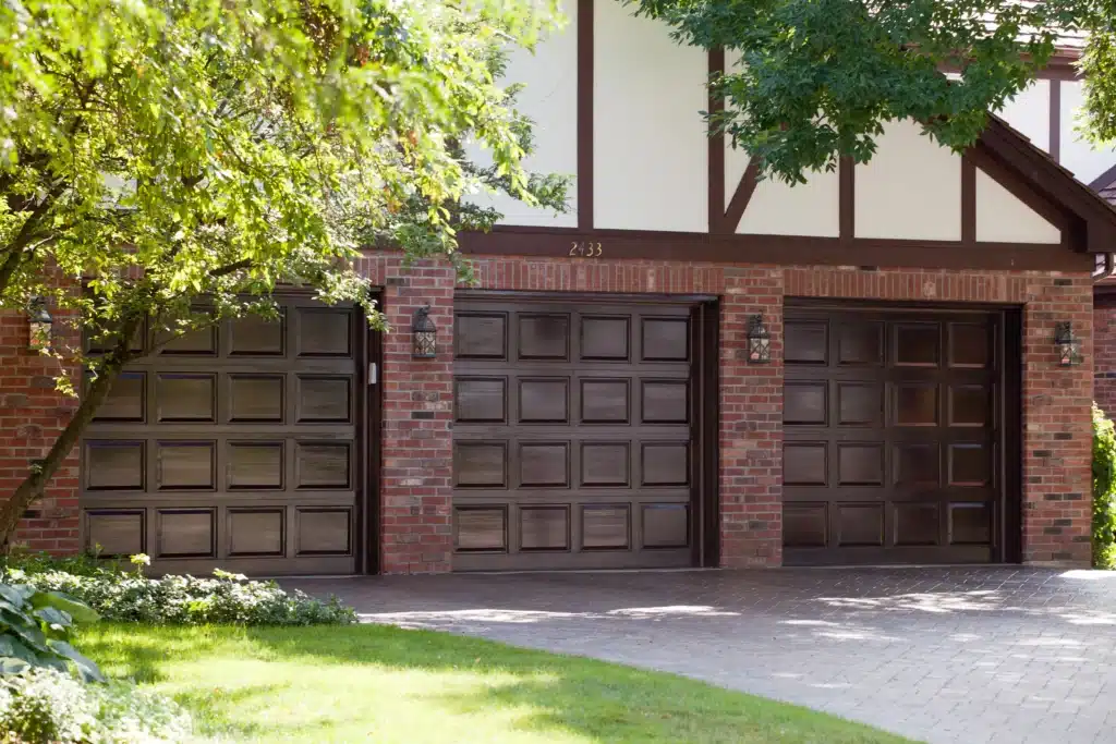Three-car garage with brick exterior and wooden doors, surrounded by trees and greenery on a sunny day.
