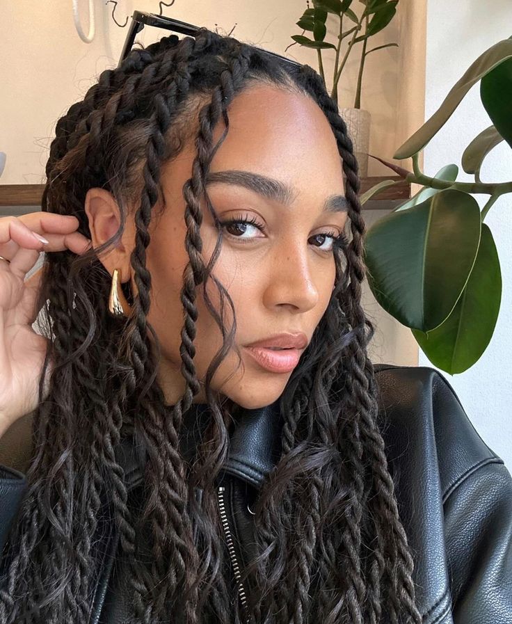 Young woman with braided hair, wearing a leather jacket, poses confidently near indoor plants.