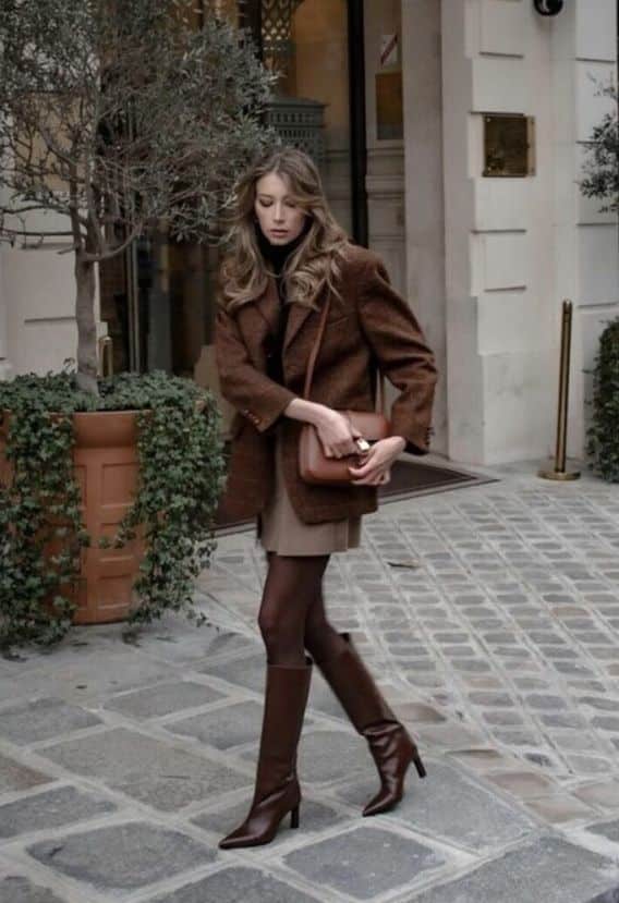 Stylish woman in brown coat and boots, walking on cobblestone street with elegant handbag, city background.