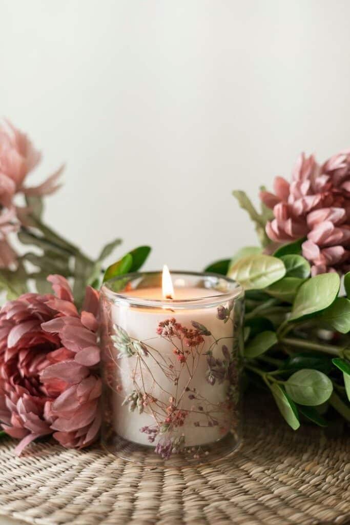 Lit floral candle with pink flowers and green leaves on wicker surface, creating a cozy, natural ambiance.