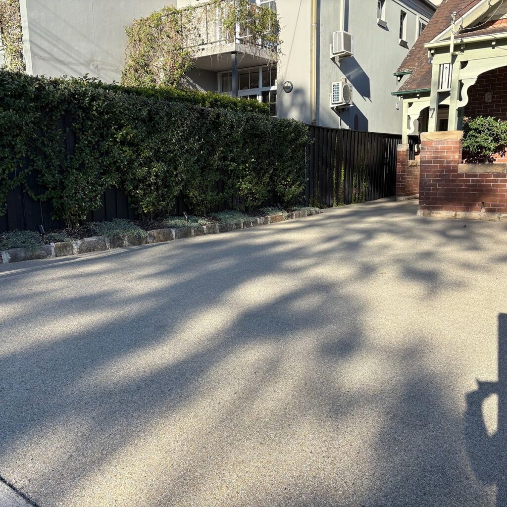 Sunny residential driveway with shadow patterns, bordered by greenery and brick walls.