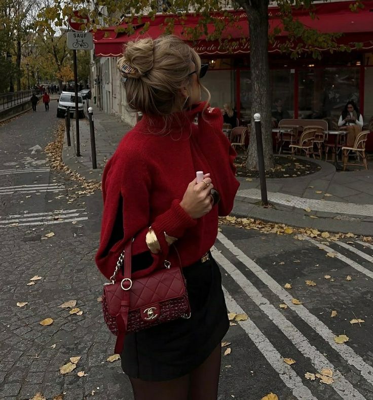 Woman in red sweater with handbag, standing on a leaf-strewn street near a French café in autumn.