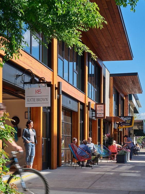 Modern shopping plaza with people relaxing outdoors, wooden architecture, and boutique shops on a sunny day.