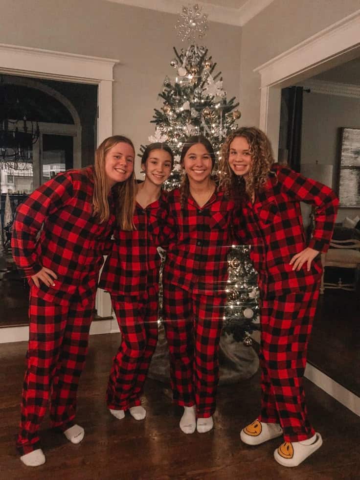 Four friends in matching red plaid pajamas pose by a decorated Christmas tree indoors.