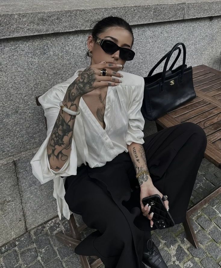 Trendy individual with sunglasses and tattoos, wearing stylish outfit, seated with designer bag on chair outdoors.