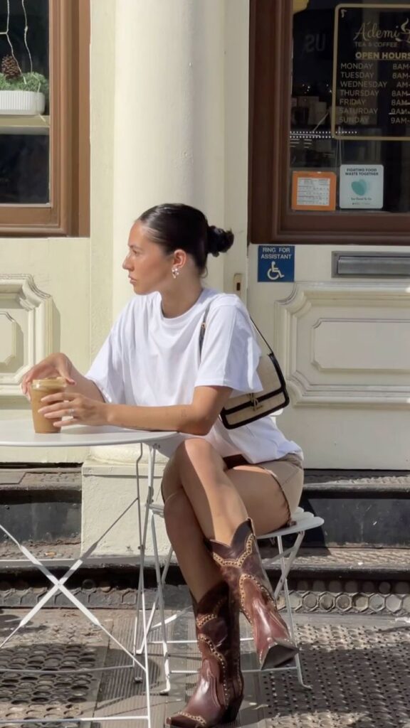 Woman in white t-shirt and boots, sitting at a cafe table with iced coffee, sunny day ambiance.