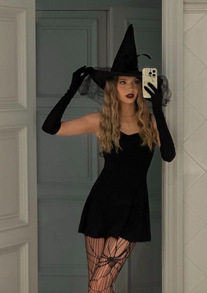 Person in a witch costume with a hat and gloves taking a mirror selfie in a stylish room.