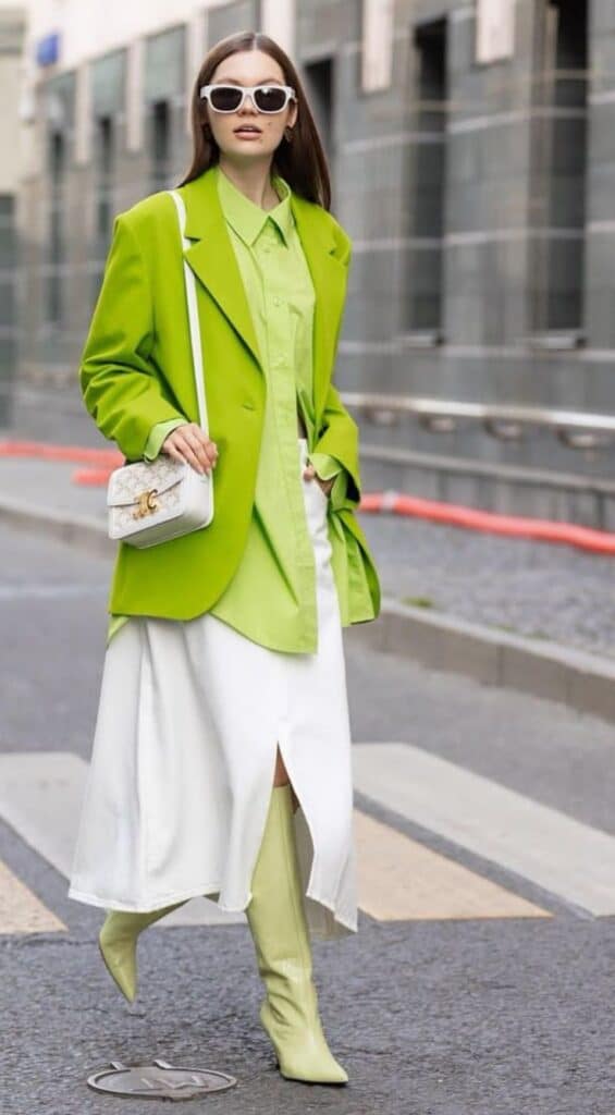 Fashionable woman in lime green blazer, white skirt, and boots, walking confidently on city street.