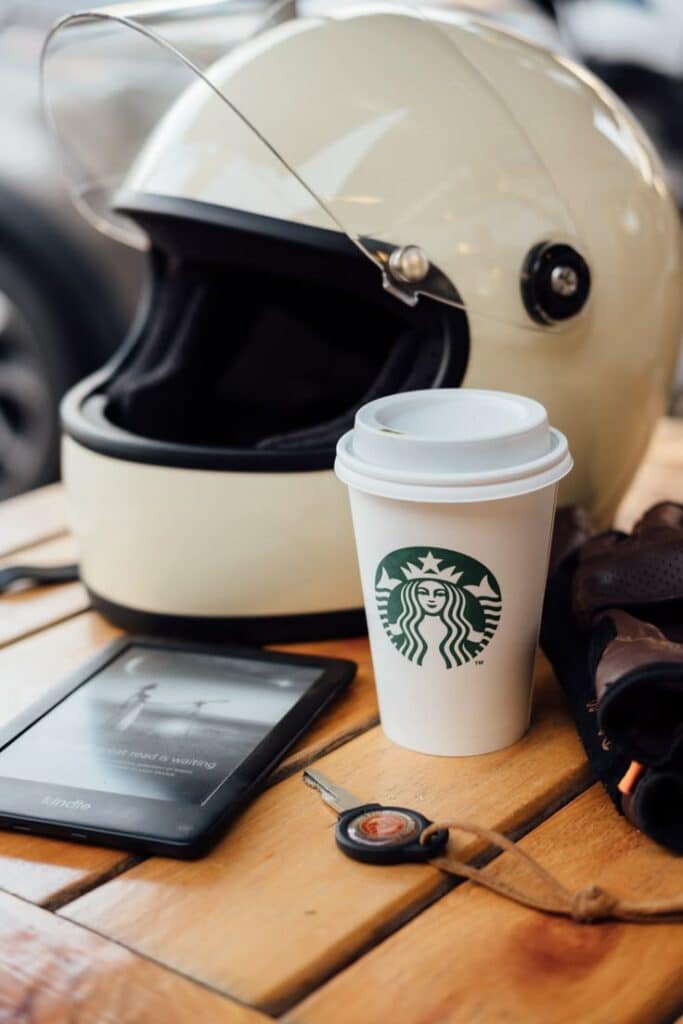 Motorcycle helmet, coffee cup, Kindle, and gloves on wooden table for a cozy break.
