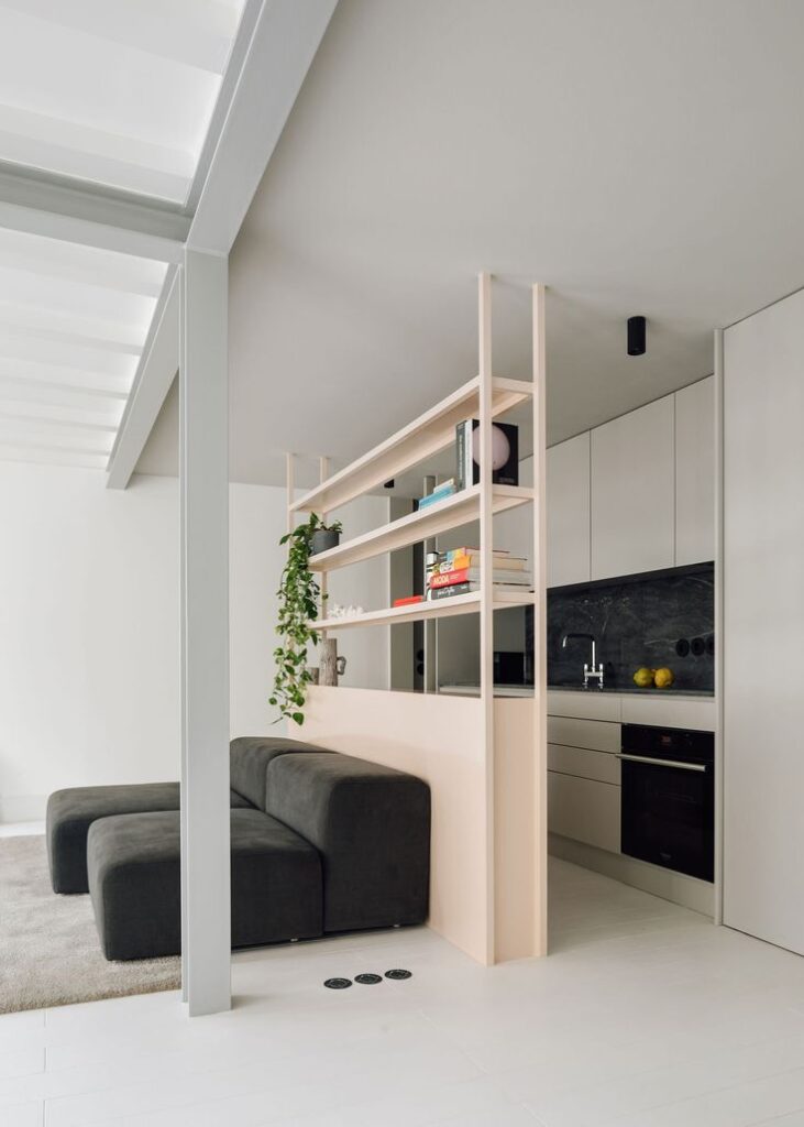 Modern open-plan living area with minimalist design, featuring a sofa, shelving, kitchen, and greenery.