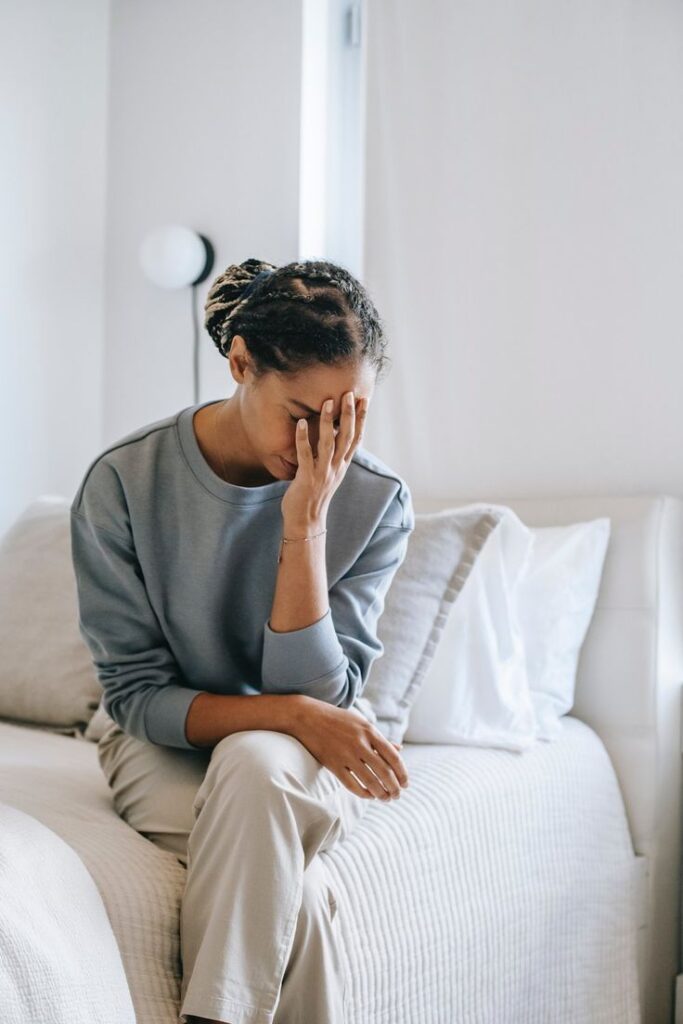 Woman sitting on bed looking stressed with hand on face, in bright minimal bedroom.