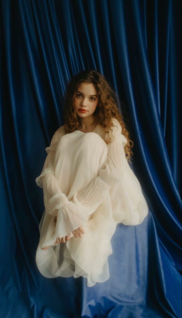 Woman in soft, white dress sits gracefully against blue velvet curtain backdrop.