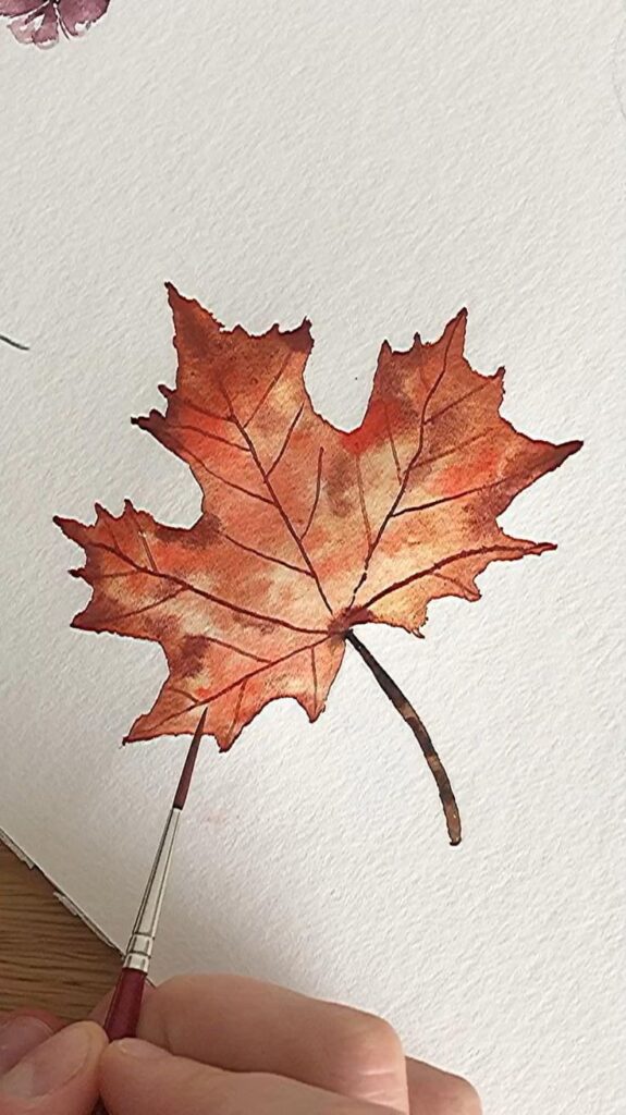 Person painting a detailed autumn maple leaf with a fine brush on white paper.