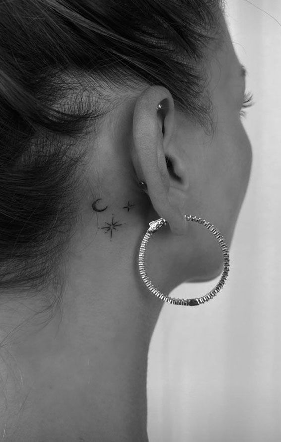 Woman with hoop earring and small moon, star tattoos behind ear in black and white.