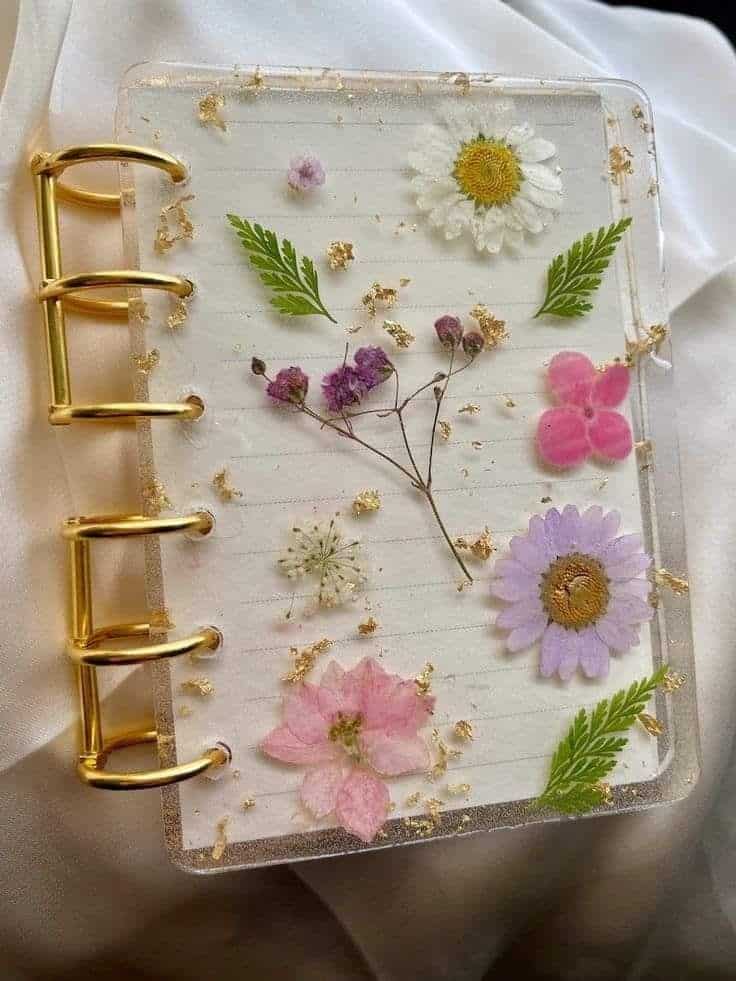 Clear notebook cover with gold rings and pressed flowers, including daisies and ferns, on a soft fabric background.