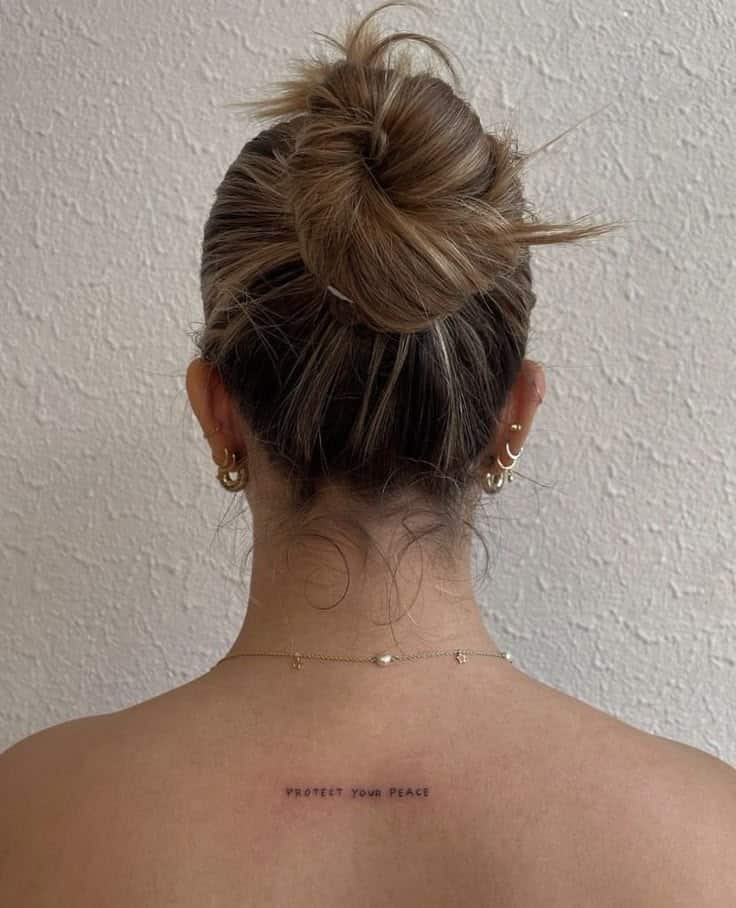 Woman with bun hairstyle, wearing multiple earrings, displays Protect Your Peace tattoo on upper back against neutral wall.