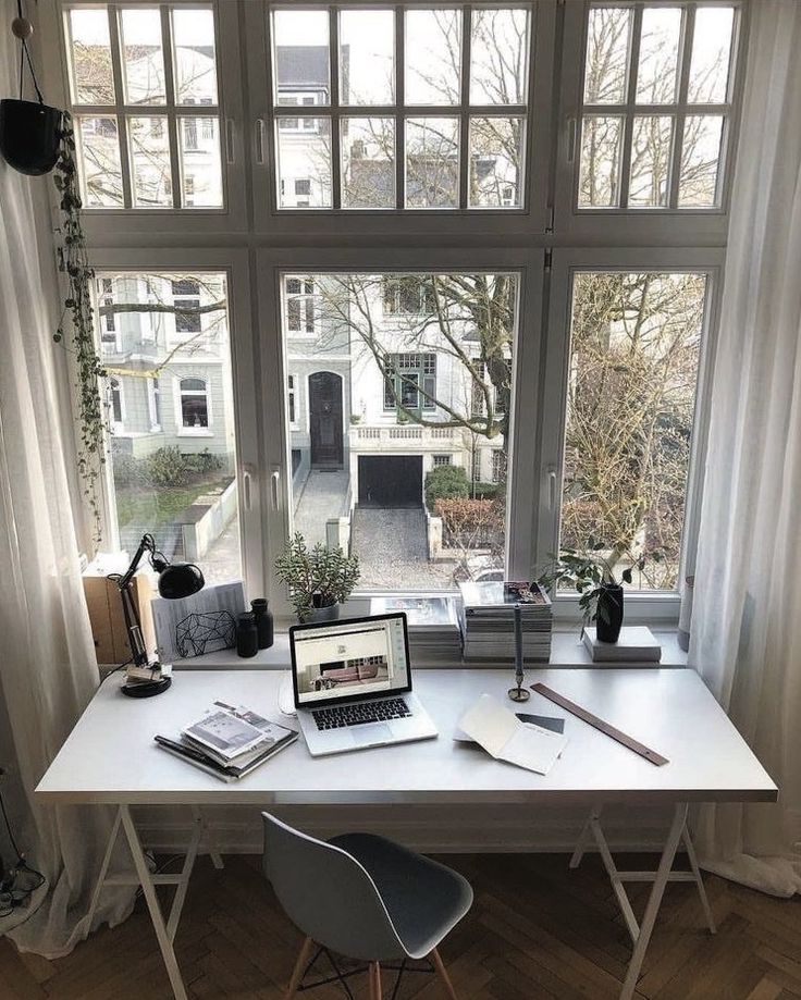 Modern home office desk by large windows with laptop, plant, and books, overlooking a residential street.