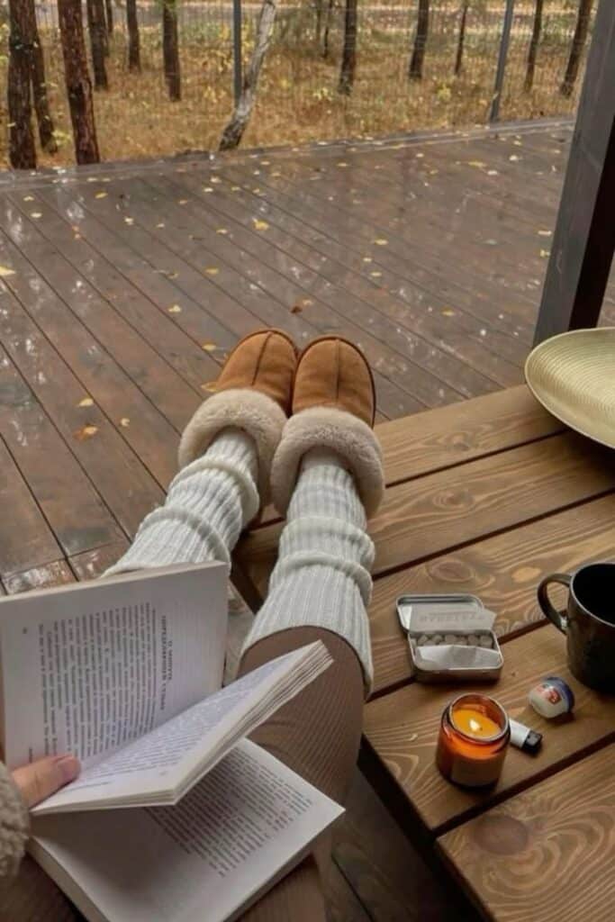 Cozy fall reading with warm slippers, book, candle, and coffee on a wooden deck in a serene forest setting.