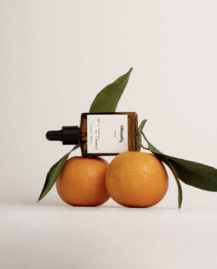 Vitamin C serum bottle with two fresh oranges and green leaves on a white background.