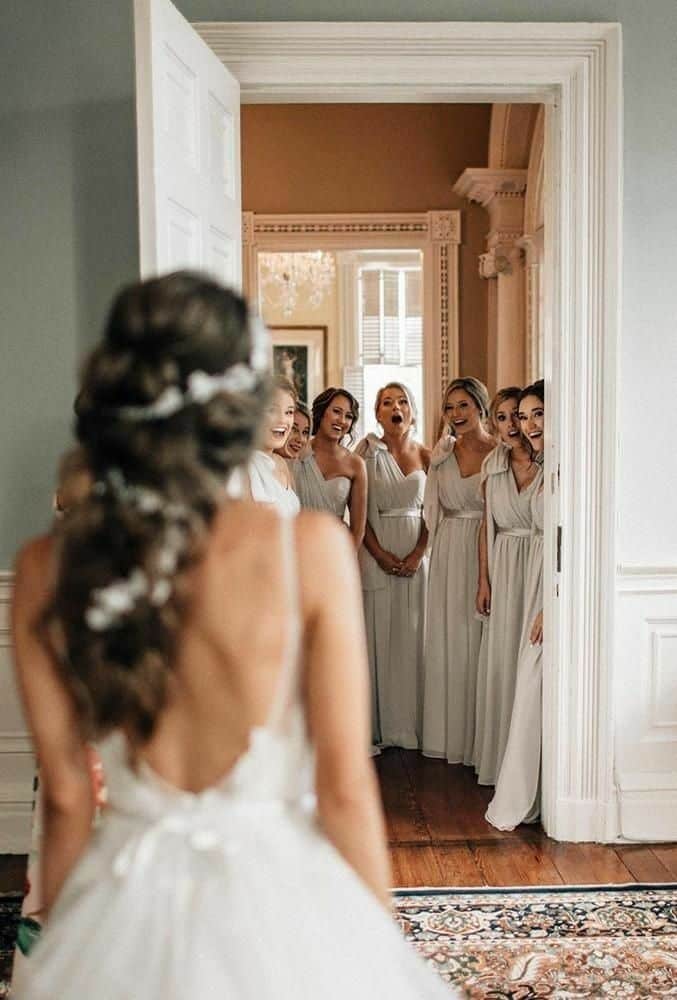 Bride reveals dress to bridesmaids, evoking surprise and joy in elegant room. Perfect bridal moment captured.