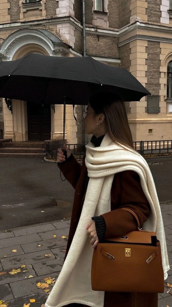 A woman holding an umbrella, wearing a stylish coat and scarf, carries a brown handbag on a rainy city street.