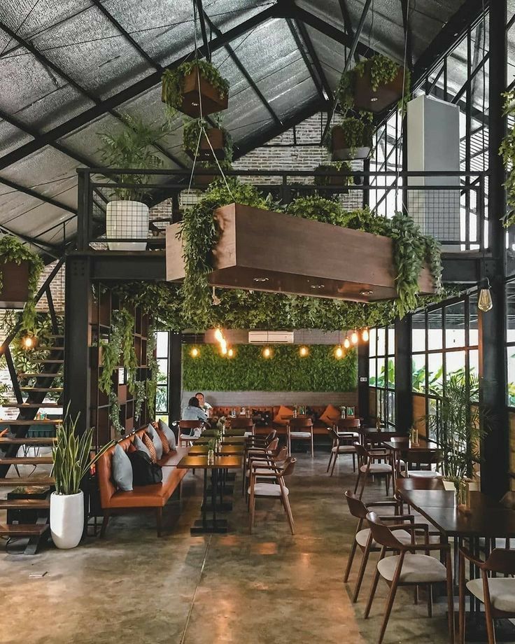 Modern café interior with greenery, hanging plants, wooden furniture, and cozy seating under industrial-style roof.