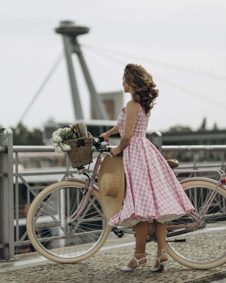 Woman in pink gingham dress with bicycle on bridge, holding hat. Scenic view with tower in background.