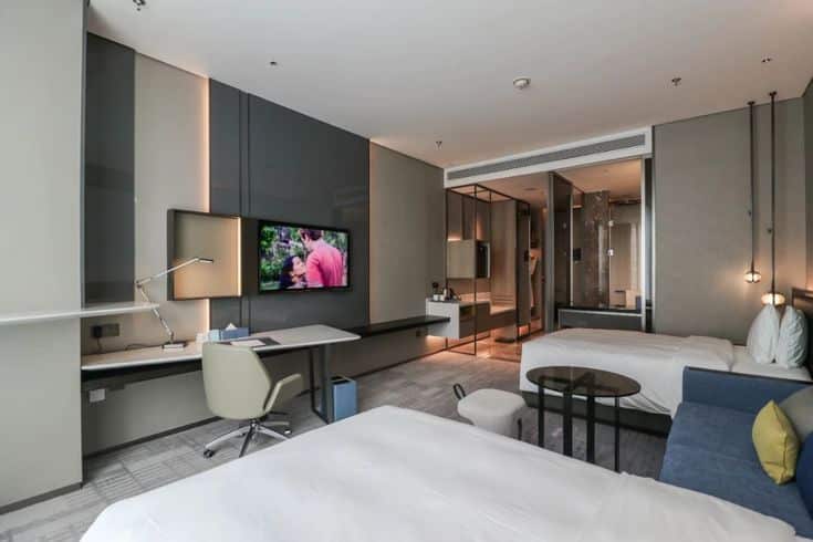 Modern hotel room with twin beds, a flat-screen TV, work desk, and glass-walled shower area for a luxurious stay.