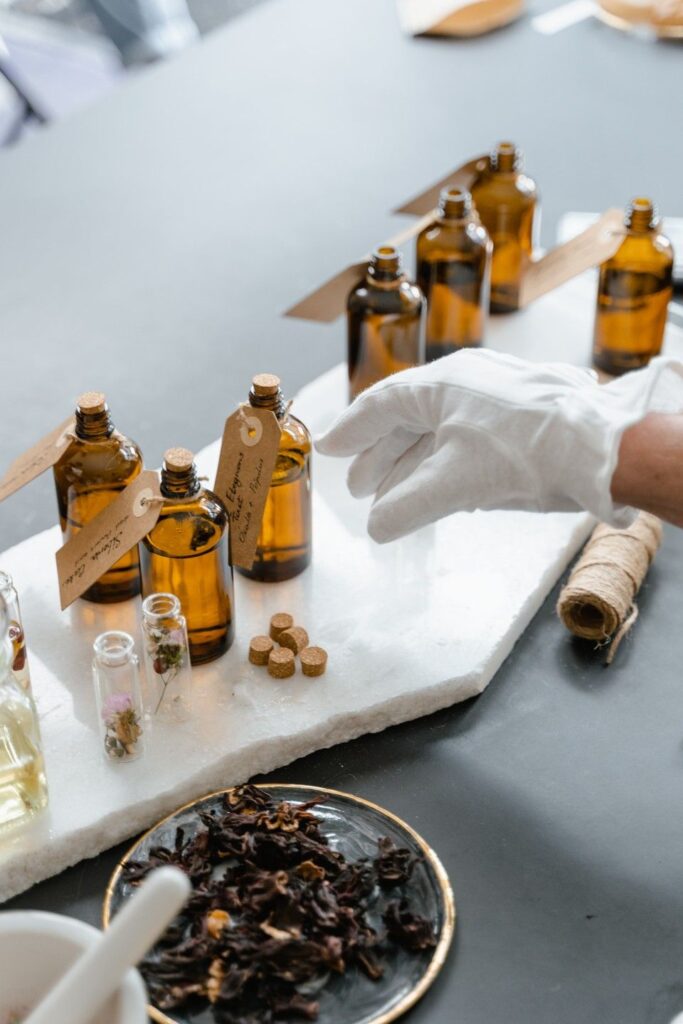 Brown glass bottles and dried herbs on a marble tray, with a hand wearing a glove, suggesting a natural apothecary setup.