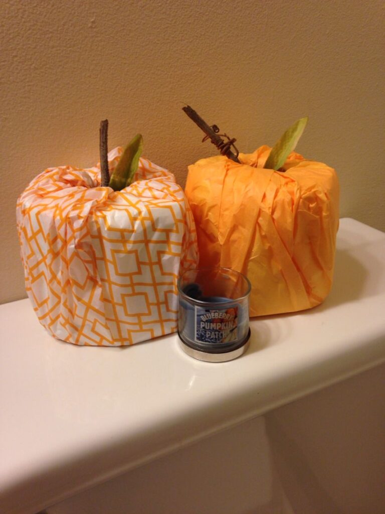 DIY toilet paper roll pumpkins with orange tissue paper and a small candle on a bathroom counter.