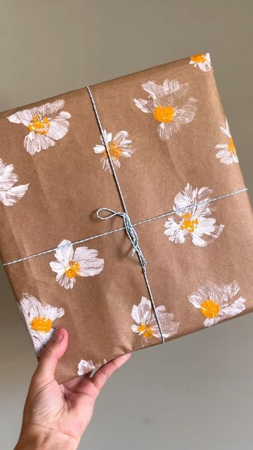 Brown gift wrapped with white flower pattern held by a hand, tied with string against a neutral background.