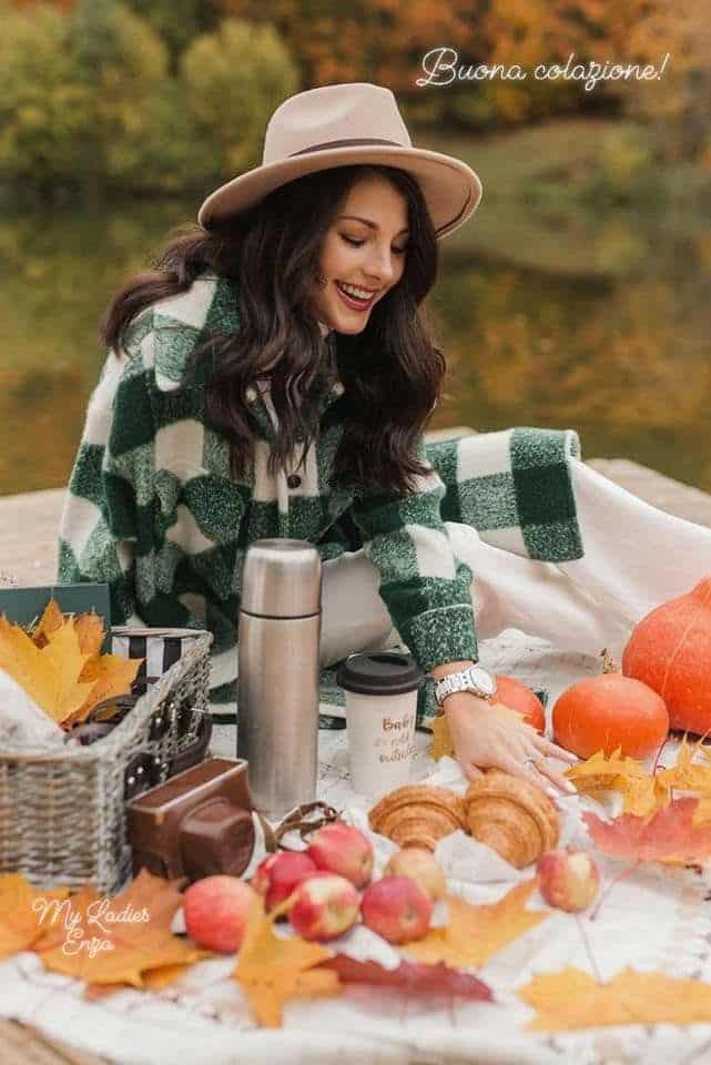 Woman enjoying autumn picnic with croissants, apples, and pumpkins by a lake, wearing a hat and green coat.