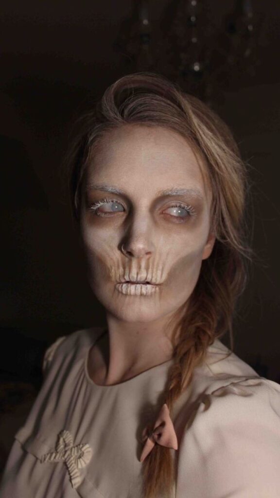 Person with skeletal makeup and braided hair, wearing a bow, gazes sideways against a dark background.