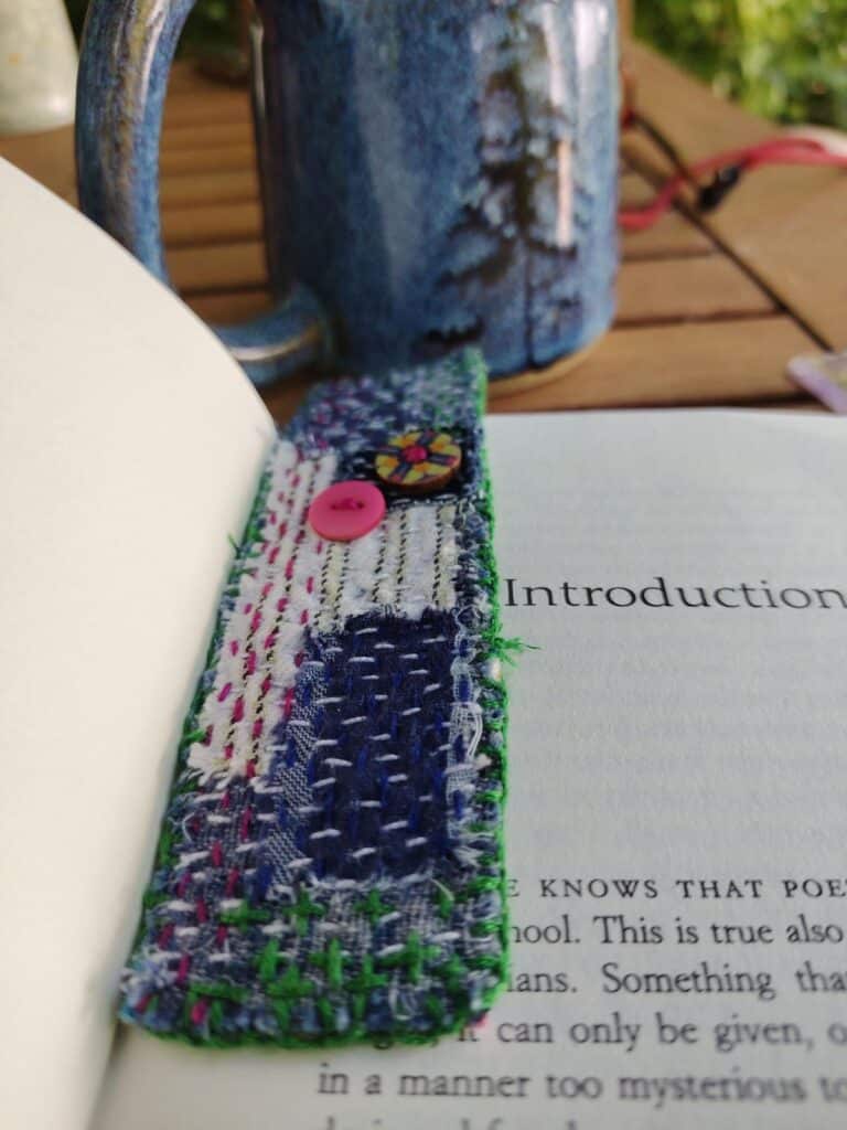 Colorful handmade fabric bookmark in book on wooden table with blue mug in background.