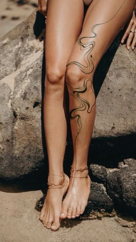 Person with leg tattoo and anklet stands on sandy beach by rocks.