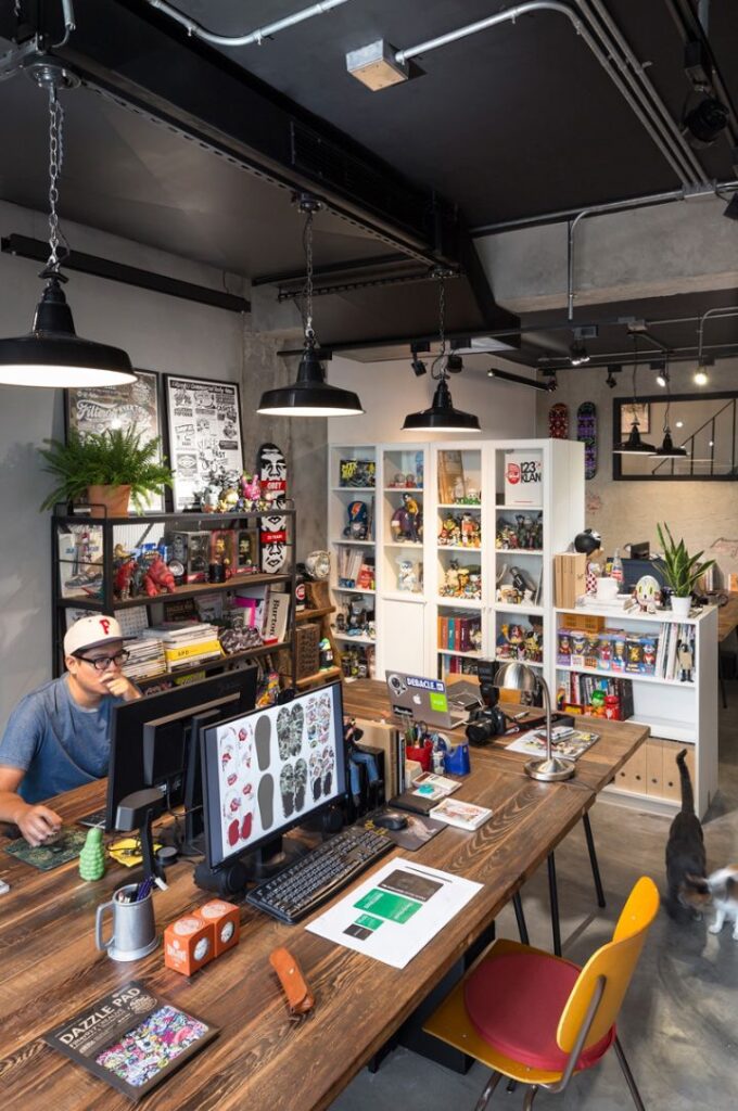 8cbb74f7-5216-4578-8538-909899ad9ba5 Creative workspace with designer at computer, colorful toys on shelves, and industrial lighting. | Sky Rye Design Creative workspace with designer at computer, colorful toys on shelves, and industrial lighting.