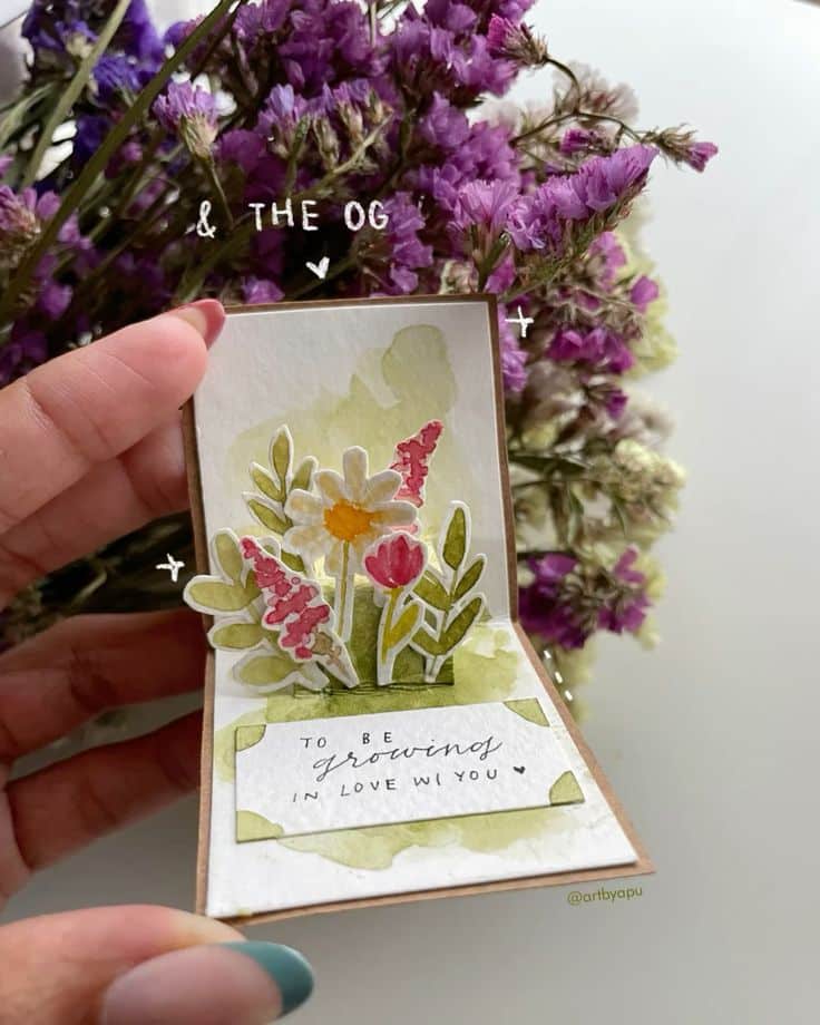 8f5a7fd96a3e2c967074115b4373e9bb Hand holding a floral pop-up card with a message about growing in love, set against a background of purple flowers. | Sky Rye Design Hand holding a floral pop-up card with a message about growing in love, set against a background of purple flowers.