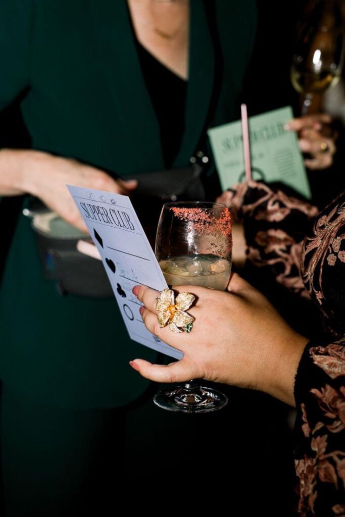 Woman holding a glittery flower ring, a cocktail, and a Supper Club menu at a social event.