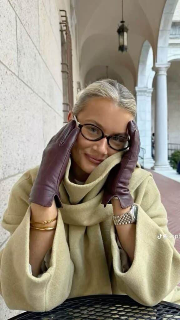 Woman in stylish coat and glasses, wearing brown gloves, smiling in an elegant outdoor setting.