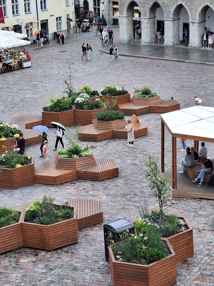 European town square with modern wooden planters, pedestrians with umbrellas, and historic stone architecture.