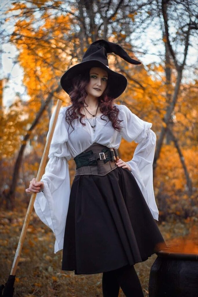 Witch in autumn forest wearing a black hat with a broomstick, surrounded by vibrant orange foliage.