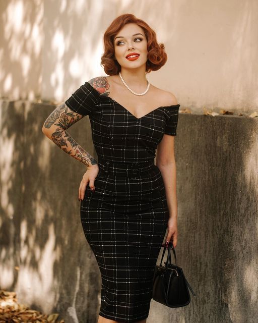 Woman in a plaid dress with tattoos and red hair stands outdoors holding a handbag, smiling confidently.