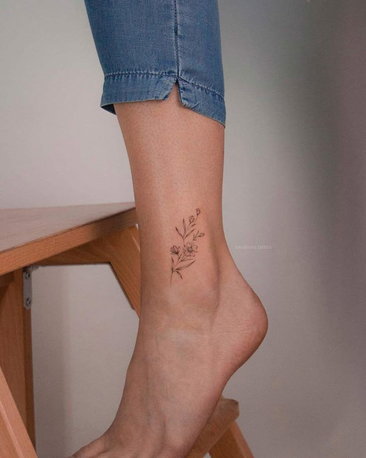 Delicate floral ankle tattoo on a person's leg wearing jeans, standing on a wooden step stool.