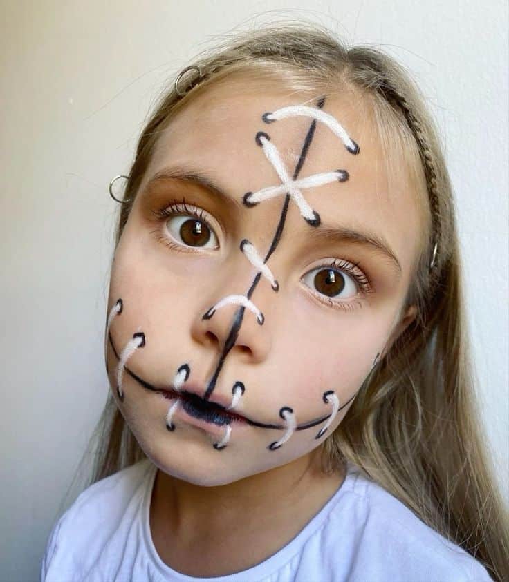 Child with creative face paint design resembling sewing stitches, wearing a white shirt, against a plain background.