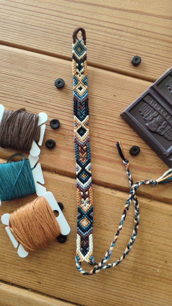 Colorful handmade friendship bracelet with geometric design, surrounded by thread spools and chocolate on wood table.